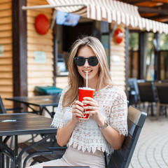 Happy fashion beautiful hipster girl with sunglasses in a fashionable lace top sits in a summer cafe and drinks cola