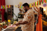 Fototapeta Na drzwi - Side view of happy Hispanic man in Mexican shirt bringing homemade snacks for festive table against his girlfriend decorating living room