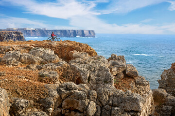 Wall Mural - happy active senior woman cycling at the the rock cliffs and lighthouse of Cabo Sao Vicente, the south-western spit of Europe at the atlantic coast of Algarve, Portugal, 