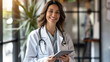 A portrait of a smiling female doctor in wearing white coat with stethoscope holding tablet and standing at modern hospital