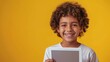 © Wendy2001 - Photo of cheerful curly little latino boy on yellow background holding tablet with toothy smile as home learning on online Internet courses for children