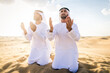 Two arab men wearing traditional emirati clothing in the desert of Dubai - Middle-eastern adult males portrait praying