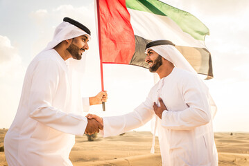Wall Mural - Two arab men wearing traditional emirati clothing in the desert of Dubai - Middle-eastern adult males portrait holding emirate flag to celebrate national day