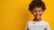 Photo of curly latino or hispanic boy on yellow background holding tablet or laptop smiling with toothy smile
