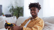 A young man in a yellow hoodie is smiling while holding a video game controller. 18 year old black boy, sitting couch in empty white room holding a gaming consol, awkward smile expression on his face