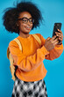 A young African American woman in casual attire takes a selfie with her cell phone while carrying a backpack, set against a blue backdrop in a studio.