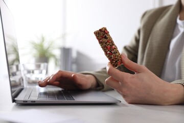 Wall Mural - Woman holding tasty granola bar working with laptop at light table in office, closeup