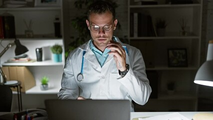 Canvas Print - Tired hispanic doctor in lab coat with stethoscope working on laptop at night in clinic office