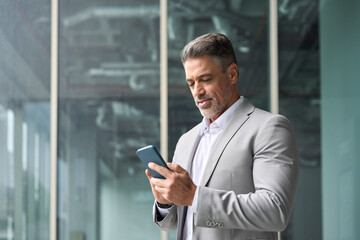 Successful rich middle aged businessman using cellphone, busy older business man leader investor, mature male executive in suit holding mobile phone standing at office window looking at smartphone.