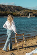 Blonde woman standing on sandy beach admiring blue lake on warm summers evening. A lady watching yachts on a lake in the sunshine. Female in jeans and white shirt poses with a chair on a lake shore..