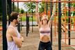 A man and a woman in sportswear workout together under a clear sky in front of a gym. The personal trainer motivates her.