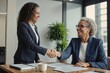 Two women shaking hands in a business setting. One woman is smiling and the other is smiling as well