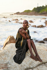 Wall Mural - Queer black person in luxury dress, jewelry sits on rocks in ocean. Lgbtq ethnic fashion model wearing jewellery dressed in posh gown poses gracefully in tropical seaside location. Pride LGBTQIA