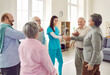 Happy young nurse at retirement home meets with group of older adults, welcomes new patients, and exchanges handshakes with old woman while other elderly people are applauding. Senior care concept