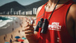 A close-up of a lifeguard's whistle and binoculars hanging around their necks, with a busy beach in the background. The words 