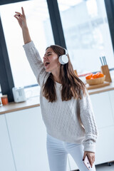 Wall Mural - Pretty young woman listening to music with mobile phone while singing and dancing in the kitchen at home.