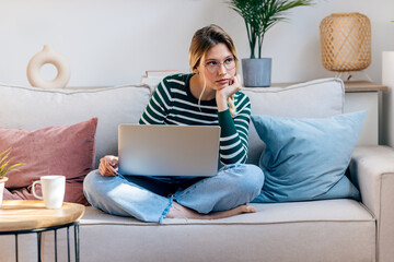 Pretty young bored woman working with her laptop while sitting on couch at home.