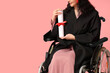 Female graduate student in wheelchair with diploma on pink background
