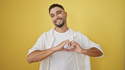 Wall Mural - Handsome young arab man making heart symbol with hands for love sign, standing isolated against yellow wall, expressing happy and romantic emotion