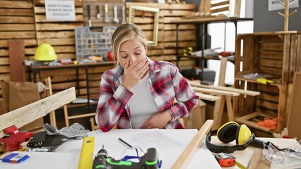 Sticker - Shocked young blonde carpenter woman, exciting surprise encounter evokes amazement and fear while sitting at carpentry table