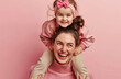 A happy mother is carrying her daughter on the shoulders, laughing and having fun together isolated over pink background with studio light in pastel colors