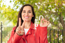 Middle Aged Woman Holding Invisible Braces At Outdoors Smiling And Showing Victory Sign