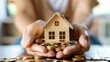 Hands holding a wooden house model over a pile of coins, symbolizing real estate investment and home savings.