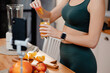 Cropped view of the fitness blogger preparing orange juice while recording video on smartphone about healthy dieting. Young woman in sport outfit standing on kitchen