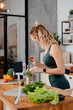 Focused blonde fitness blogger preparing lemonade while recording video on smartphone about healthy dieting. Young woman in sport outfit standing on kitchen