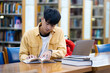 A young man is sitting at a table in a library, reading a book