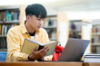 A young man is sitting at a desk with a laptop and a book