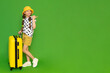 A little girl with a suitcase on wheels is looking at your advertisement. A child in shorts and a hat is resting for the summer. Green isolated background. Copy space.