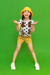A little girl in a hat and shorts is enjoying the warm summer. Children's summer holidays.  The young girl spread her palms in surprise and smiles broadly. Green isolated background.
