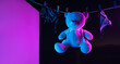 A toy teddy bear in a bdsm mask and leather straps hangs on a clothesline in neon light on a dark background