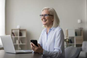 Canvas Print - Overjoyed mature businesswoman laugh on message received seated at desk at modern workplace, get great news, having pleasant on-line remote chat, enjoy new mobile application. Tech, workflow, business