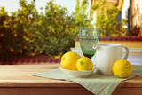 Fototapeta  - Mediterranean summer concept.  Lemons and white jug on wooden picnic table over lemon tree background