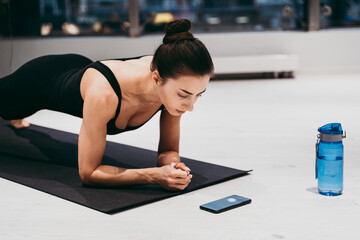 Beautiful girl training in the gym. Young woman performs an abdominal plank while checking the run on the smartphone. Russian classic dancer doing stretching and yoga poses