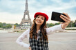 Beautiful young woman visiting paris and the eiffel tower. Parisian girl with red hat and fashionable clothes taking a pov selfie with the smartphone camera and smiling.