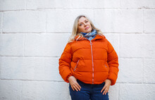 Happy Positive Middle-aged Woman In Orange Down Jacket Smiling At Camera While Posing Against Concrete Grey Wall