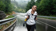 Happy young woman with backpack walking on wet road and enjoying beautiful view on mountains. Foggy and rainy weather outdoors. Active time spending.