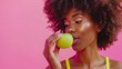 Beautiful black curly haired happy woman in bright yellow top biting a crunchy green apple carried in her hand isolated on pink background, with copy space