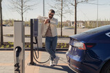 Fototapeta  - A stylish adult male checks his watch as he waits by his electric car, which is charging at a modern charging station on a sunny day.
