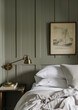 A bedroom with white linen bedding, brass wall sconces and vintage oil lamps, a wooden bedside table, and a framed drawing of sailing boats on the green wood-paneled walls, in soft neutral tones.