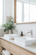 A modern minimalist bathroom with two white rectangular sinks, one on top of the other and a wood countertop, a large mirror above, an eucalyptus plant in a vase, and light oak wooden cabinets below.
