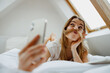 Smiling woman on bed, taking selfie with phone in comfort