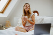 A woman with a beautiful hairstyle is sitting on the bed, smiling with a cup of coffee and a laptop