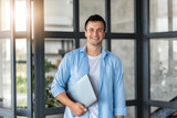Fototapeta Kuchnia - Young successful Caucasian man entrepreneur or an office worker stands with laptop in a modern office, looking at the camera and smiling