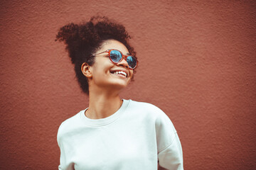 Wall Mural - African ethnicity young woman in stylish sunglasses, with curly hair tied up in high ponytail, looking away while smiling broadly showing straight perfect teeth