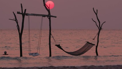 Wall Mural - Sea sunset landscape with swing and hammock on background of red sun in sky. Concept of tourism and beach vacation at resort.