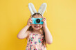 A cute little girl with bunny ears is holding an Easter egg camera and taking pictures against a yellow background with an Easter theme